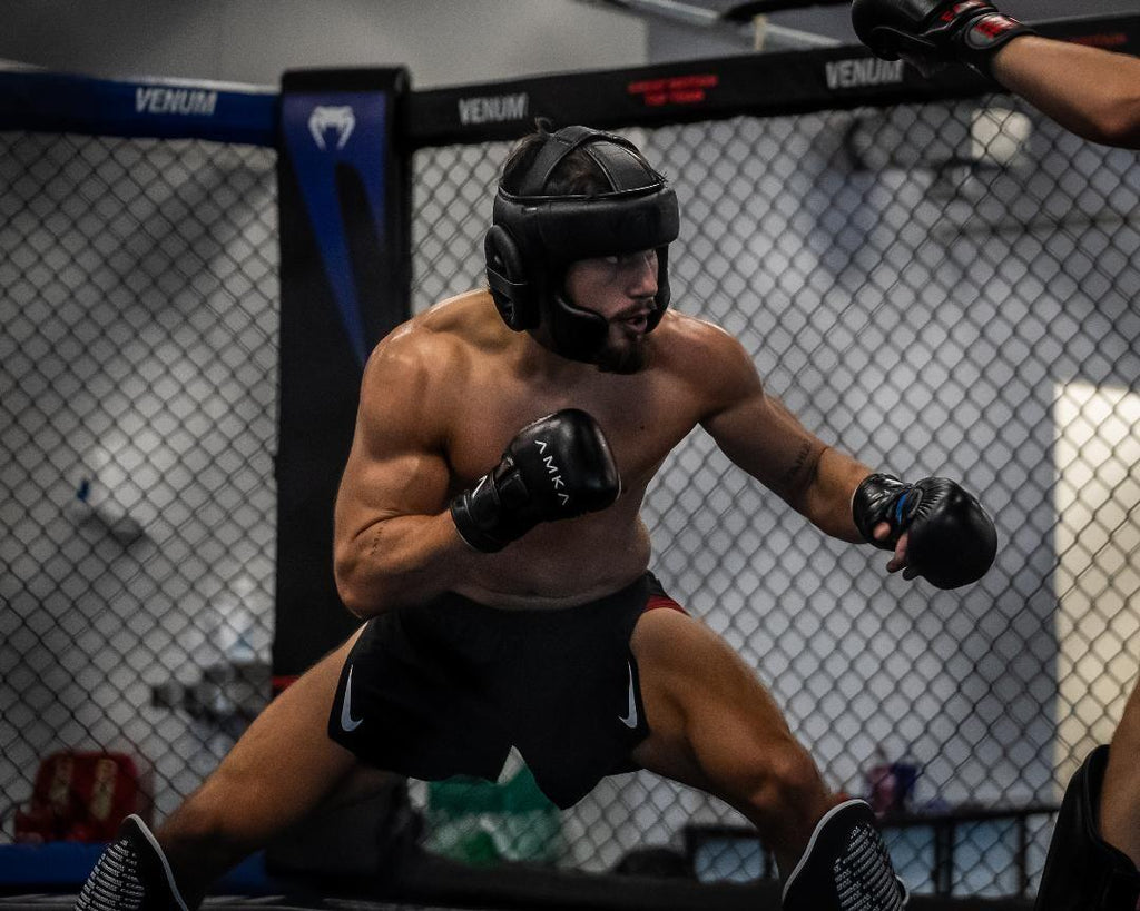 A male MMA fighter in a caged gym wearing a black headguard, black gloves, and black shorts with a white Nike swoosh, captured mid-action inside a Venum cage.

